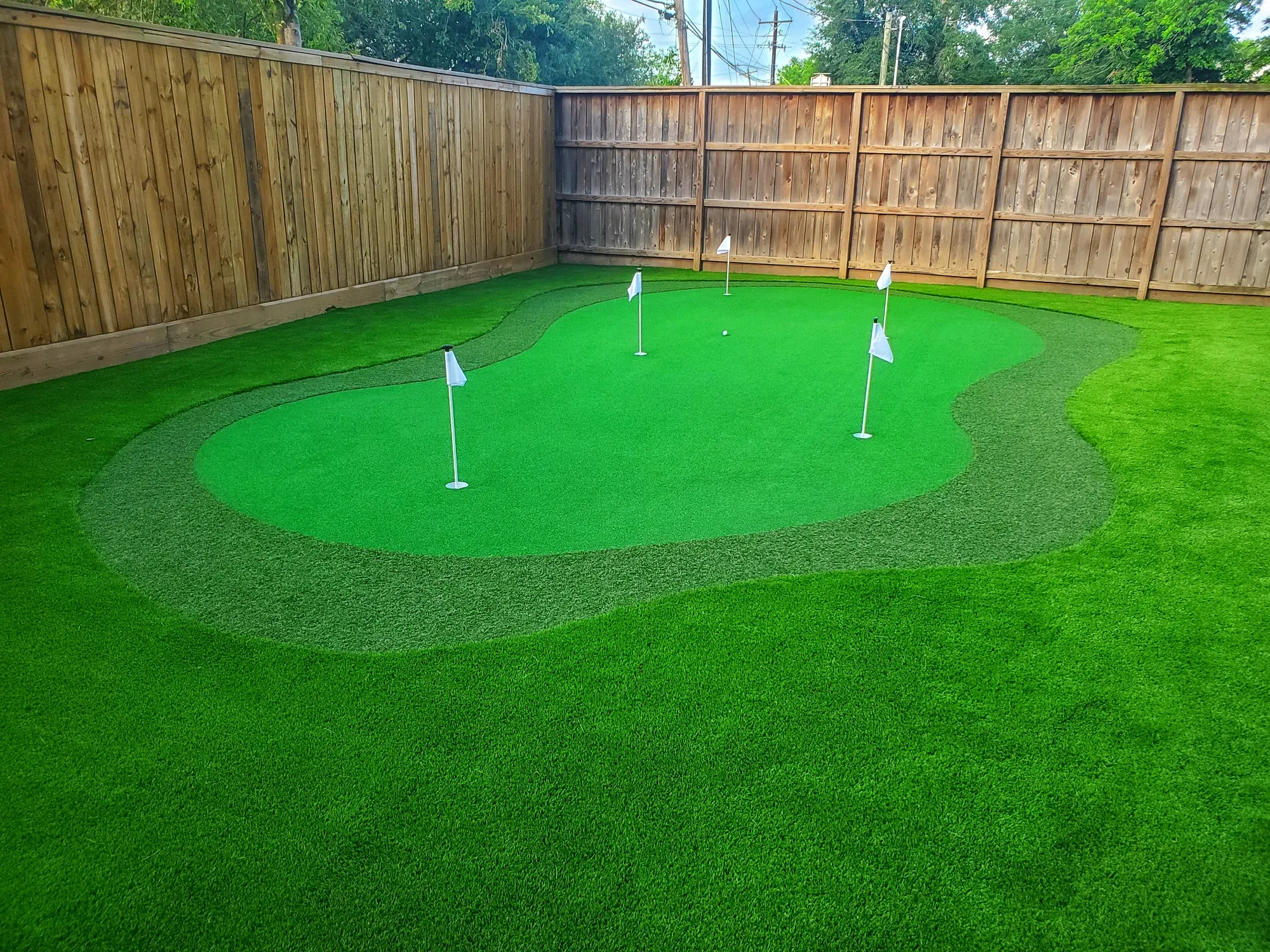 A kidney-shaped practice green with four flags set into a fenced artificial turf back lawn