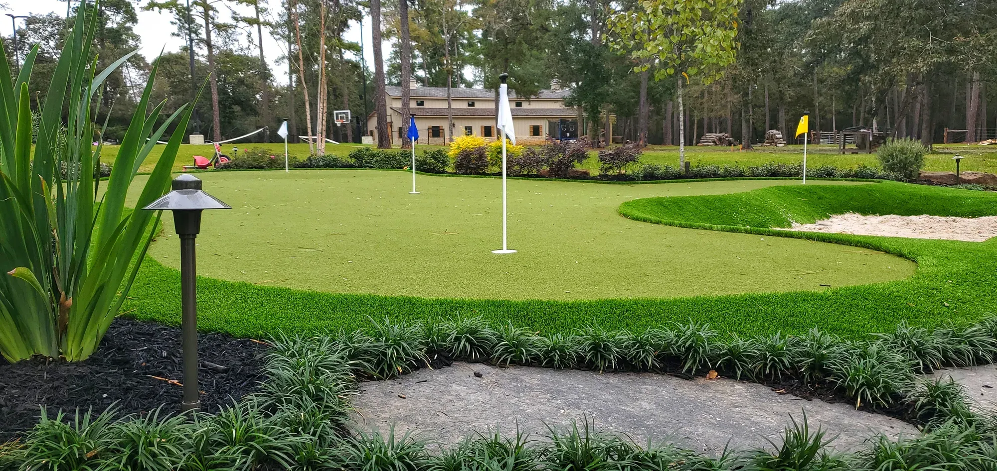 Large artificial putting green with four flags and a sand bunker cut into the fringe, in a wooded back yard