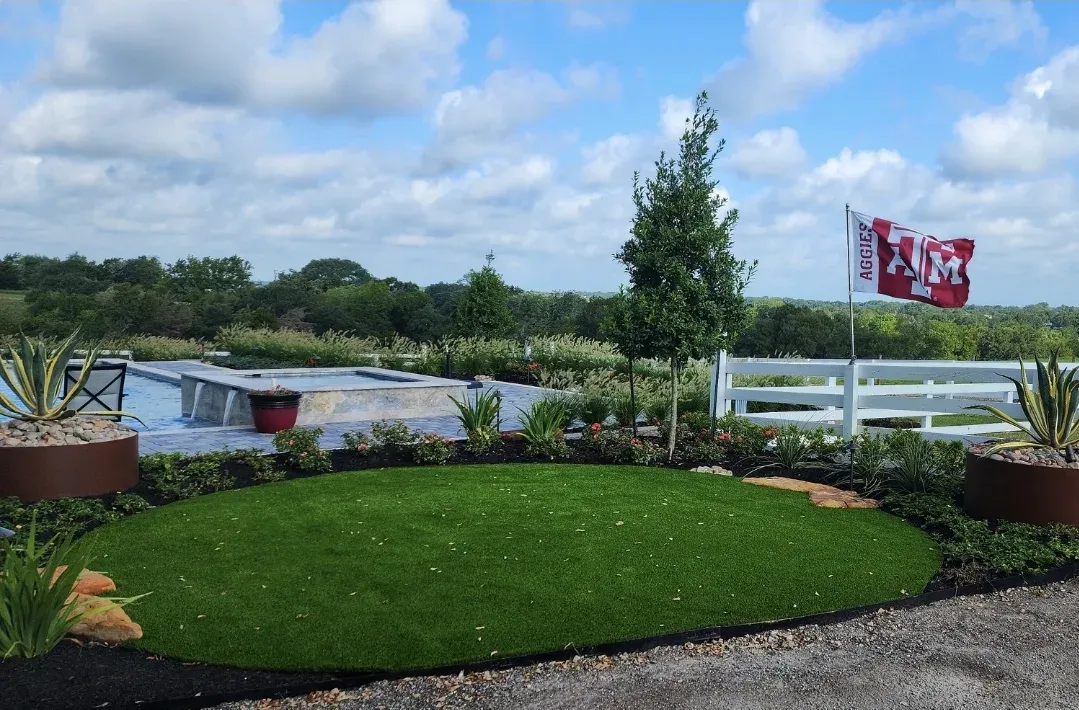 An oval artificial lawn with a paver edge, white ranch fencing and open pasture beyond