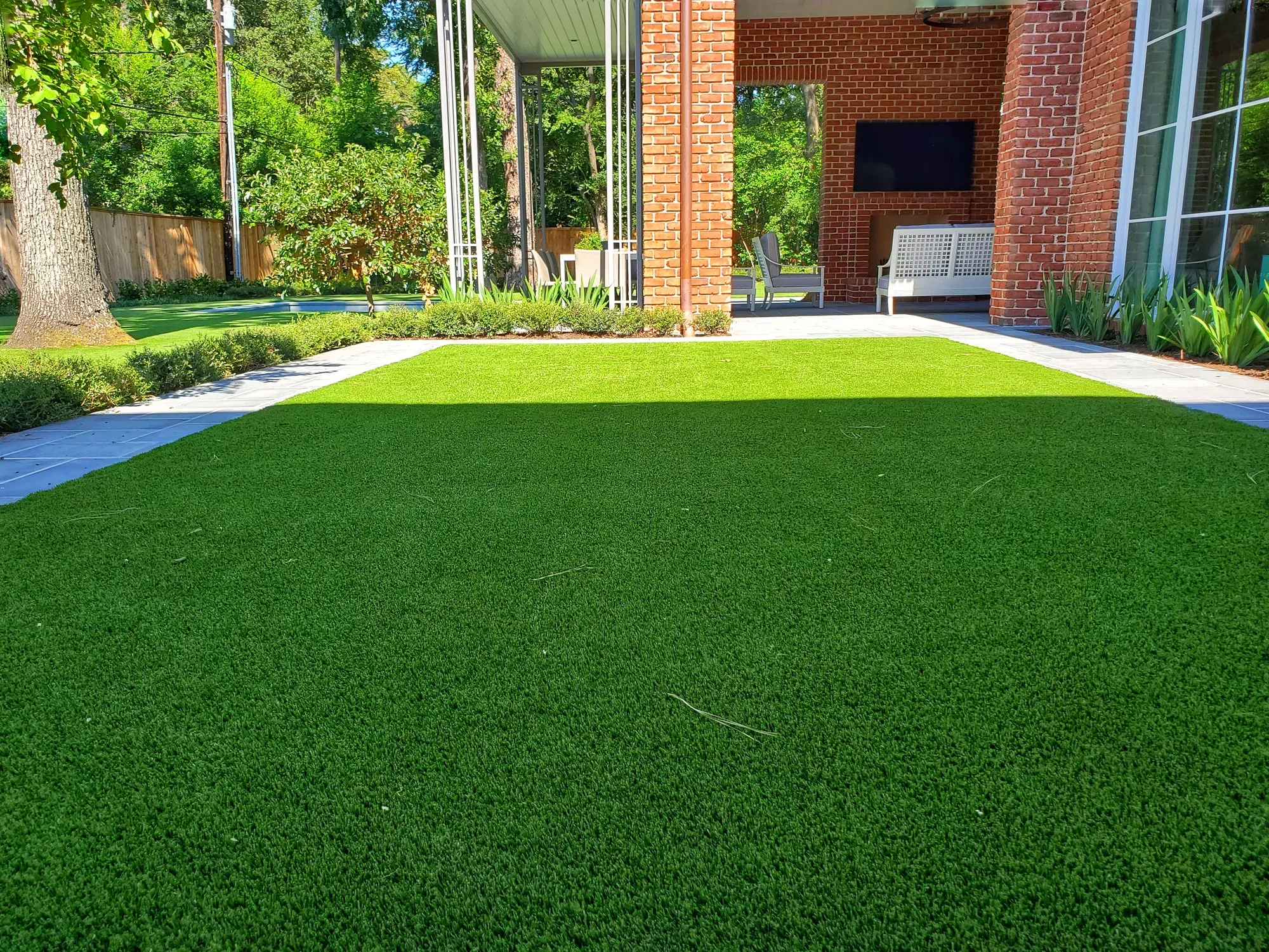 Artificial turf back lawn edged with stone paving, behind a brick house with a covered patio
