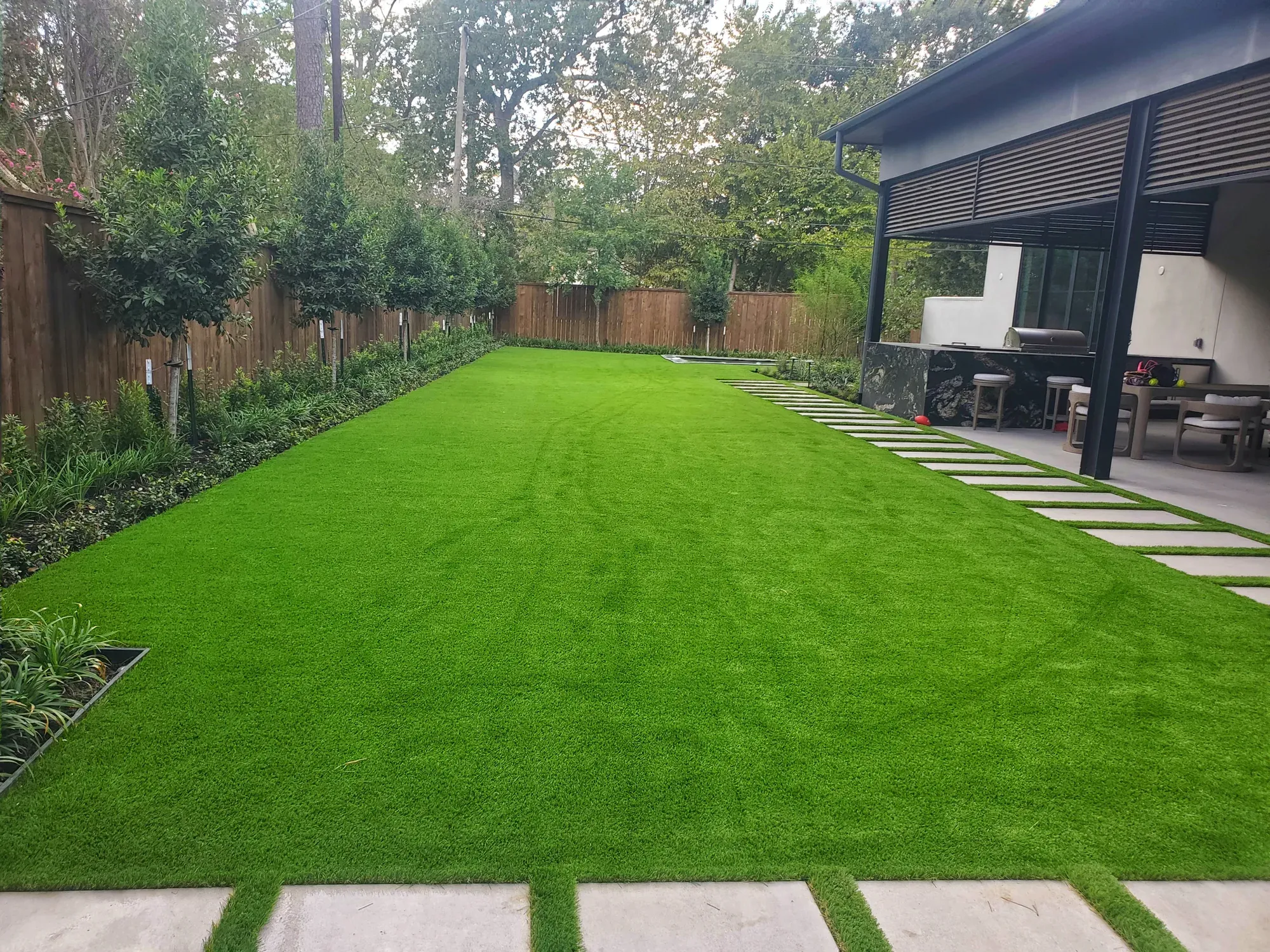 Long artificial turf back lawn running past a covered patio, edged with stone pavers and young trees
