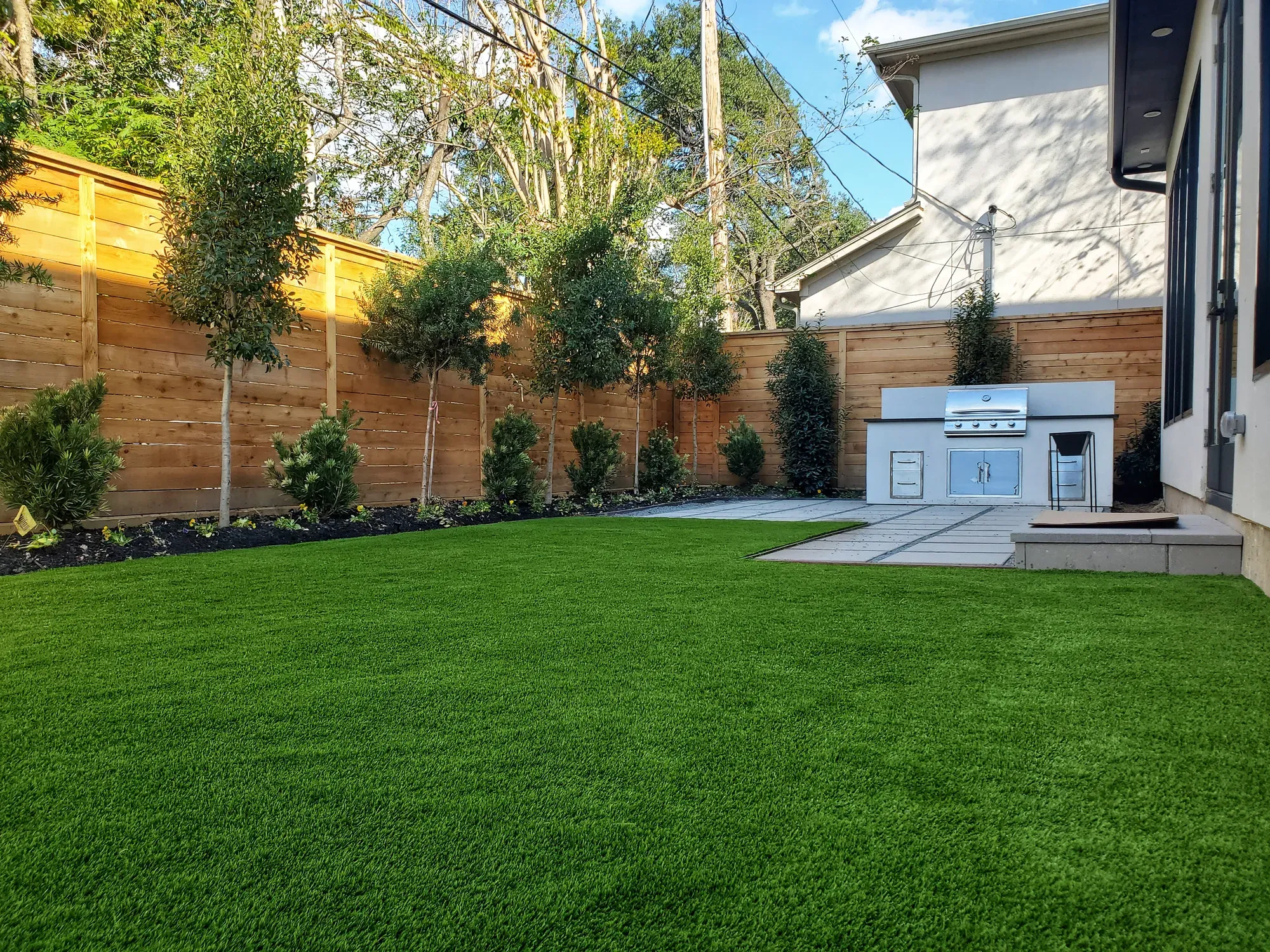 Artificial turf back lawn running up to a paved outdoor kitchen, with a cedar fence and clipped hedges behind