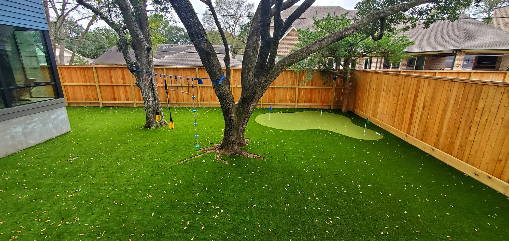 A shaded back lawn of artificial turf under a large tree, with a small practice green beside the trunk