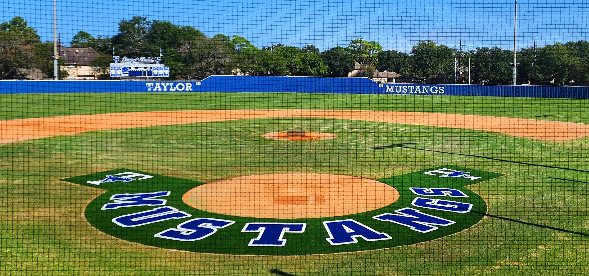 A baseball infield with a cut-in turf team logo in the halo behind home plate, seen through the backstop netting