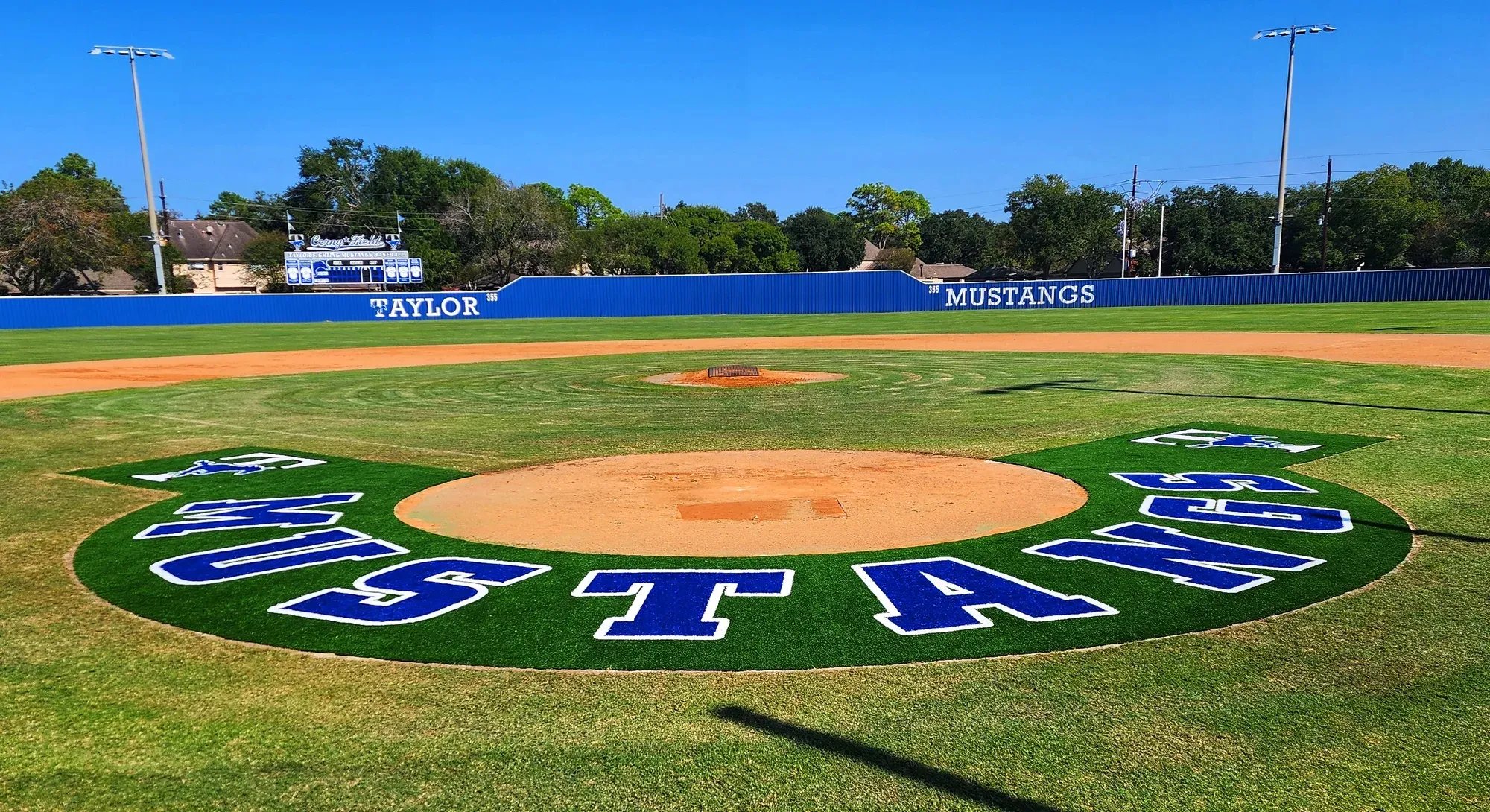 School baseball infield with the team name cut into artificial turf in a ring around the pitching mound, outfield wall and scoreboard behind