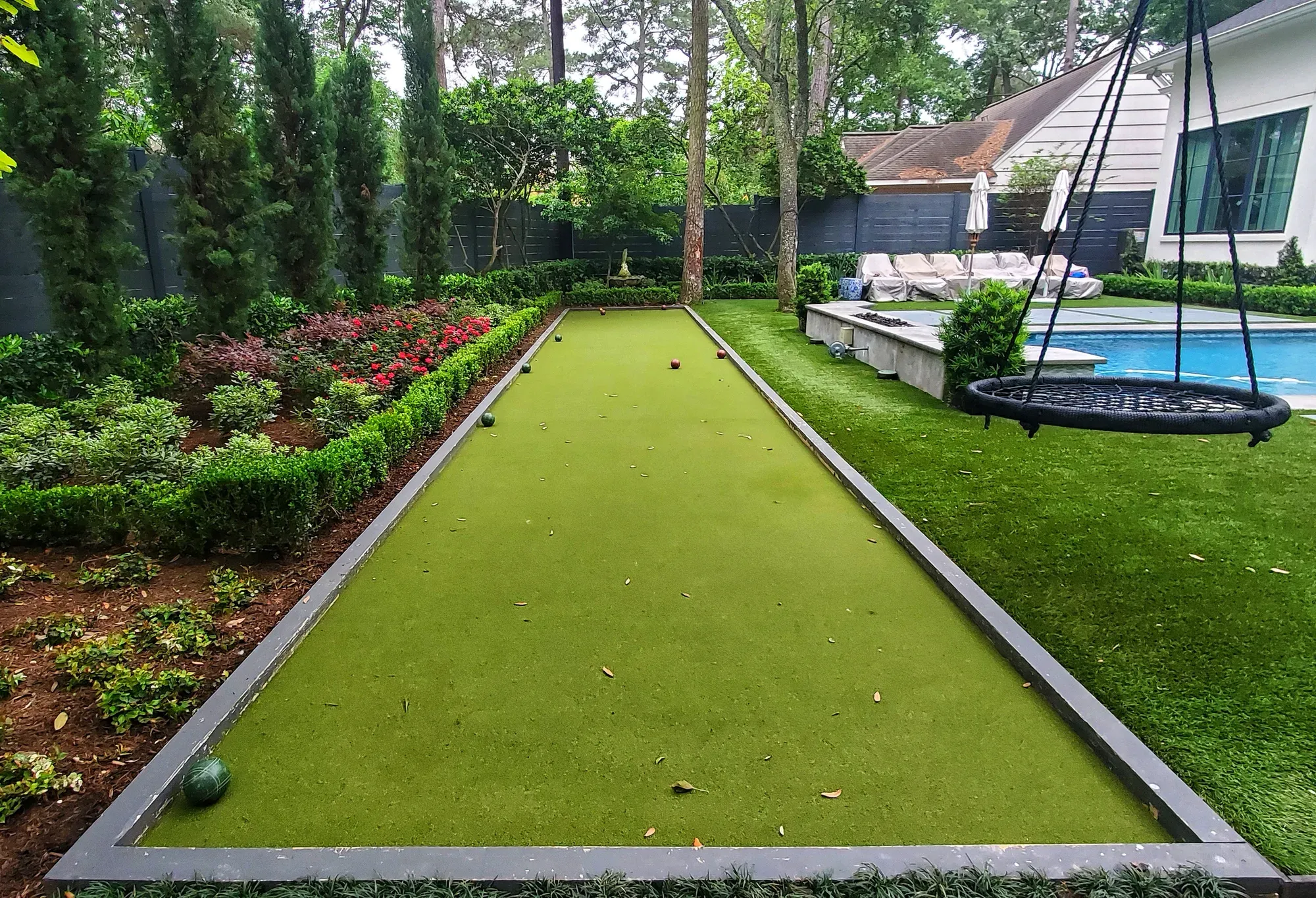 A long artificial turf bocce lane with hard edges, set in a planted garden with a hanging swing