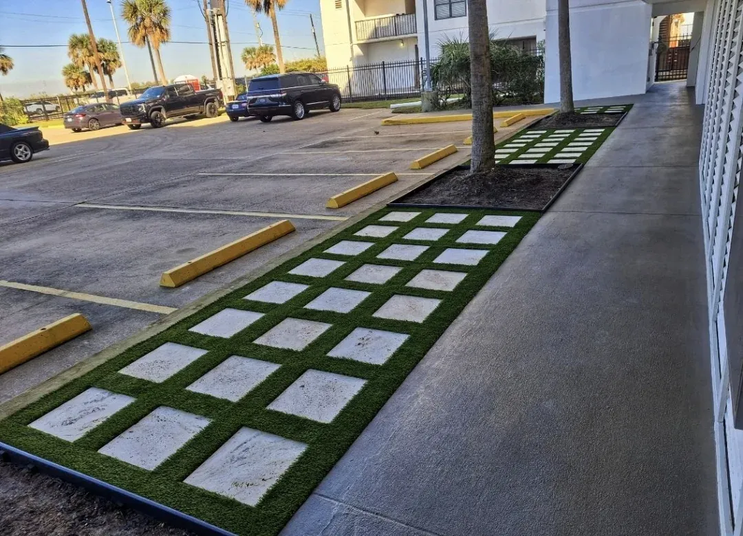 A sidewalk of alternating stone tiles and artificial turf panels beside a parking lot and palms