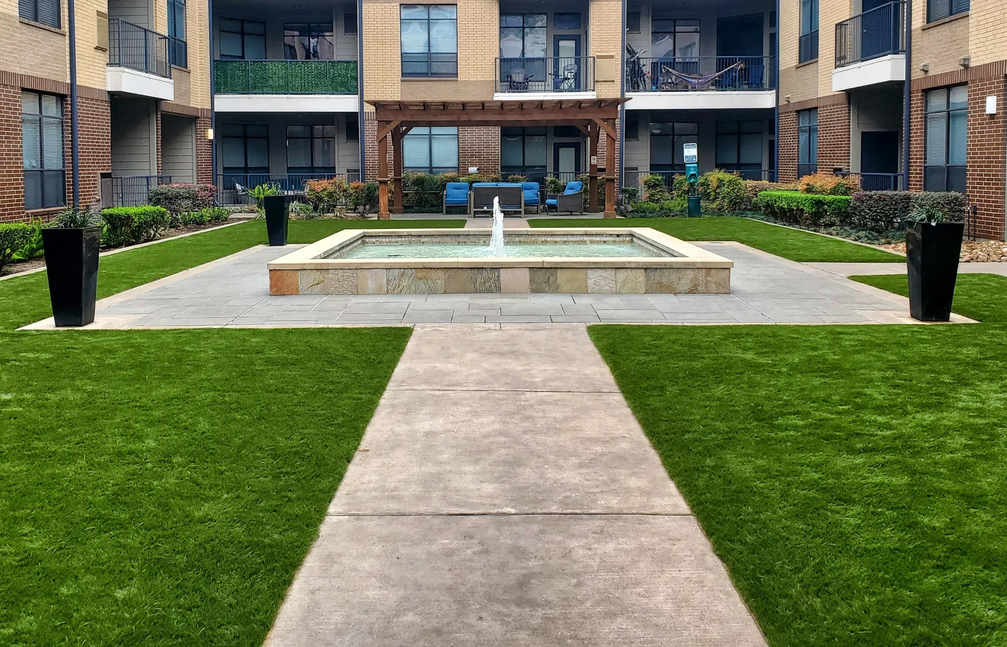 Artificial turf lawns either side of a concrete walk in an apartment courtyard, leading to a fountain and a timber pergola with seating