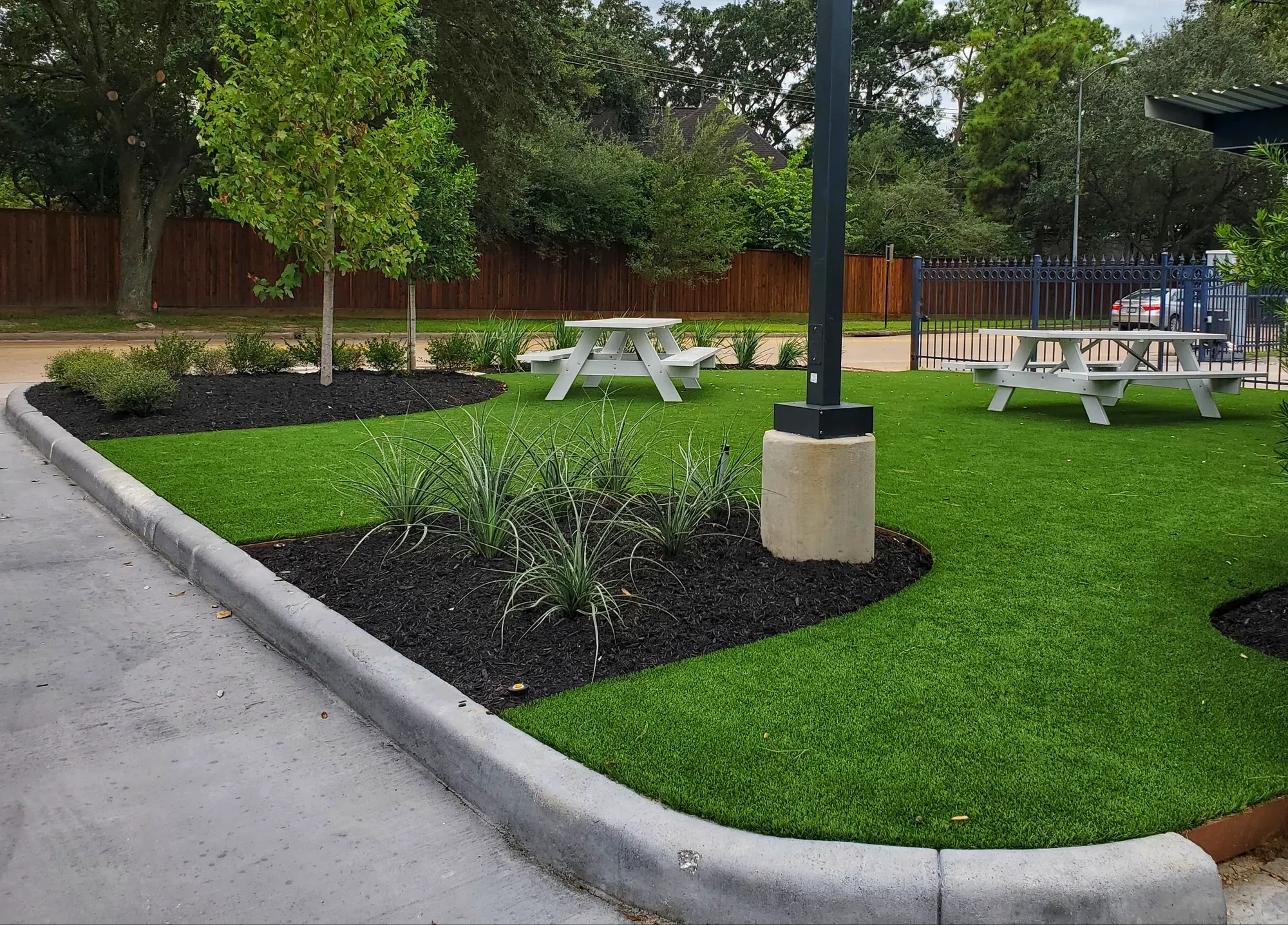 A kerbed artificial turf island with picnic tables under a shade canopy and planted beds