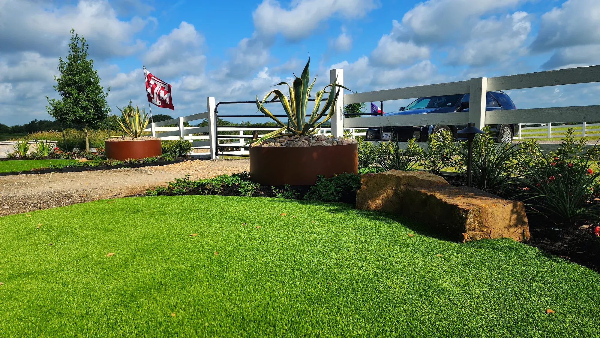 Artificial turf lawn at a commercial property entrance, edged with corten planters, ornamental beds and a white rail fence along the drive