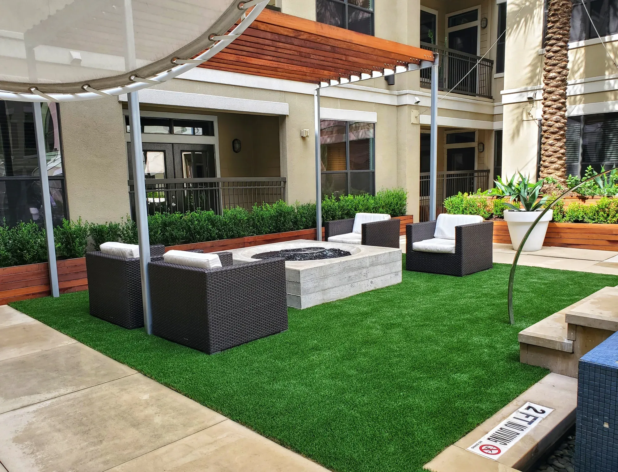 Shaded apartment courtyard with lounge chairs and a gas fire pit standing on artificial turf, planter walls around the edge