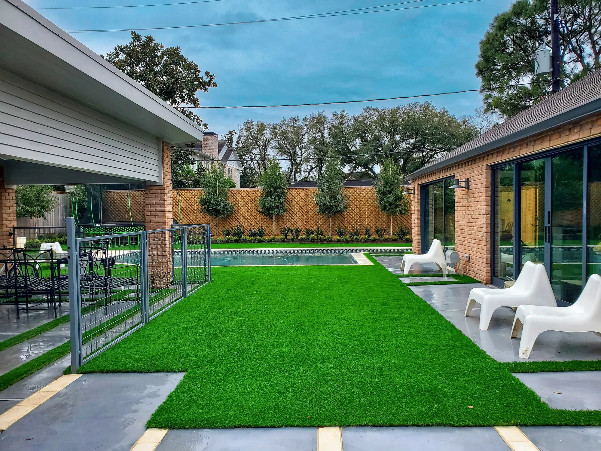 A turf lawn between a paved terrace and a fenced pool, with loungers on the grass