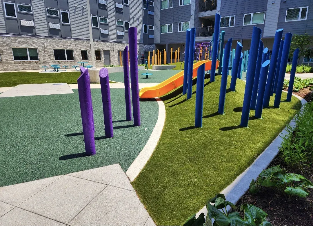 Commercial playground where artificial turf meets a poured-rubber play surface, with coloured play posts, a slide and an apartment building behind