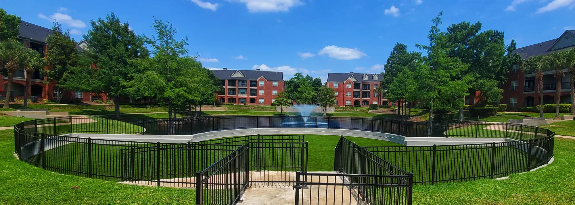 Wide artificial turf lawn wrapping a fountain pond, fenced along the water, with apartment buildings and trees behind it