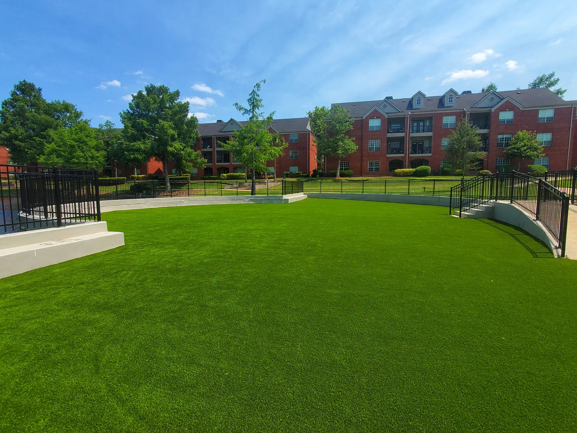A large open artificial turf lawn in front of a brick multi-family building, enclosed by black fencing