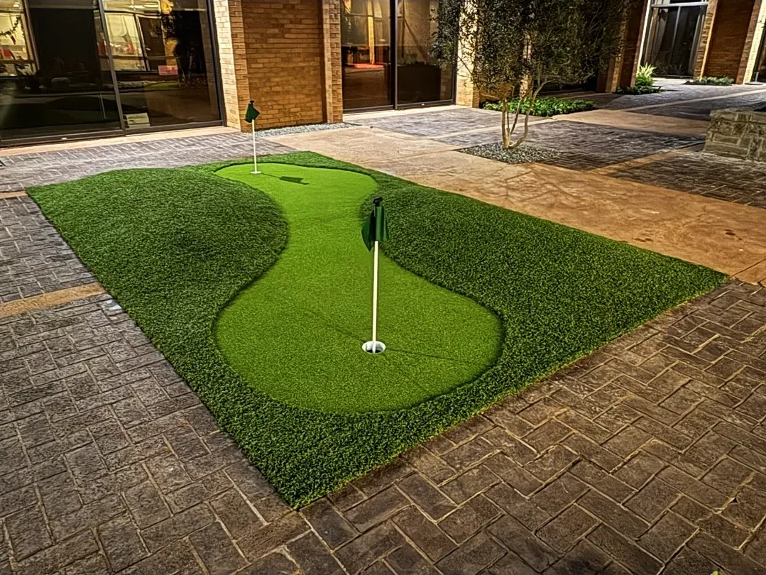 Small artificial putting green with two flagsticks, set into the paved courtyard of a brick office building