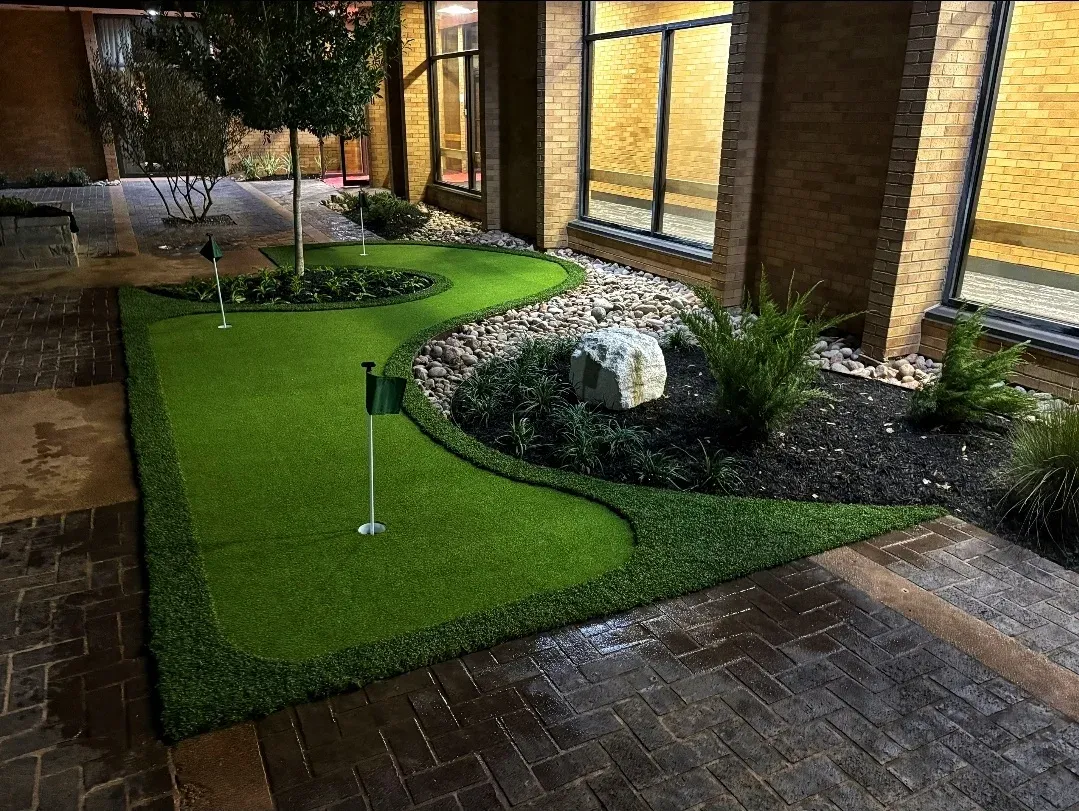 Artificial putting green with two flagsticks in a brick courtyard, lit at night with wet paving around it