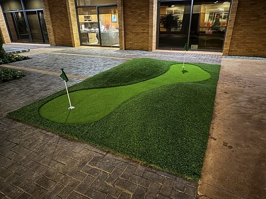 Two-tier artificial putting green with two pin flags, set into stamped concrete paving outside a lit commercial building at night