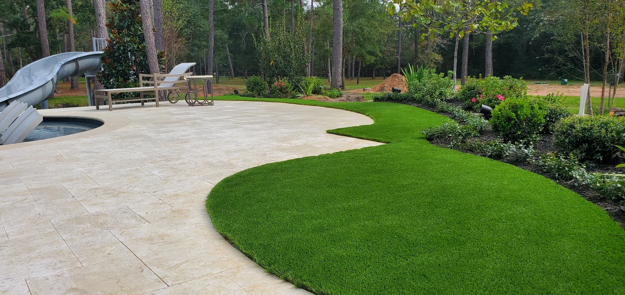 Curved artificial turf lawn meeting a travertine pool deck, with a water slide and tall pines behind