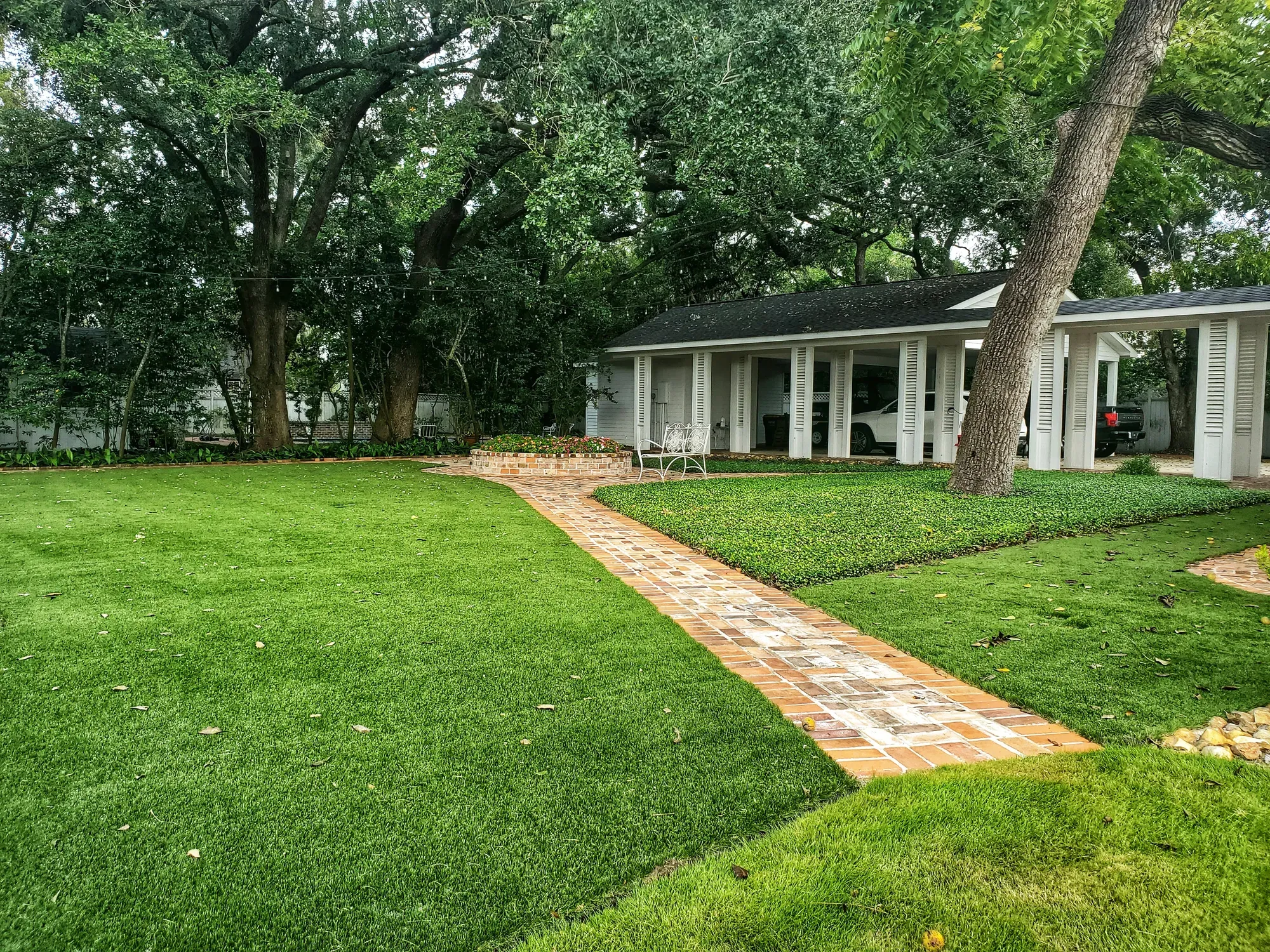Front lawn of artificial turf under mature live oaks, with a brick walkway leading to a white house