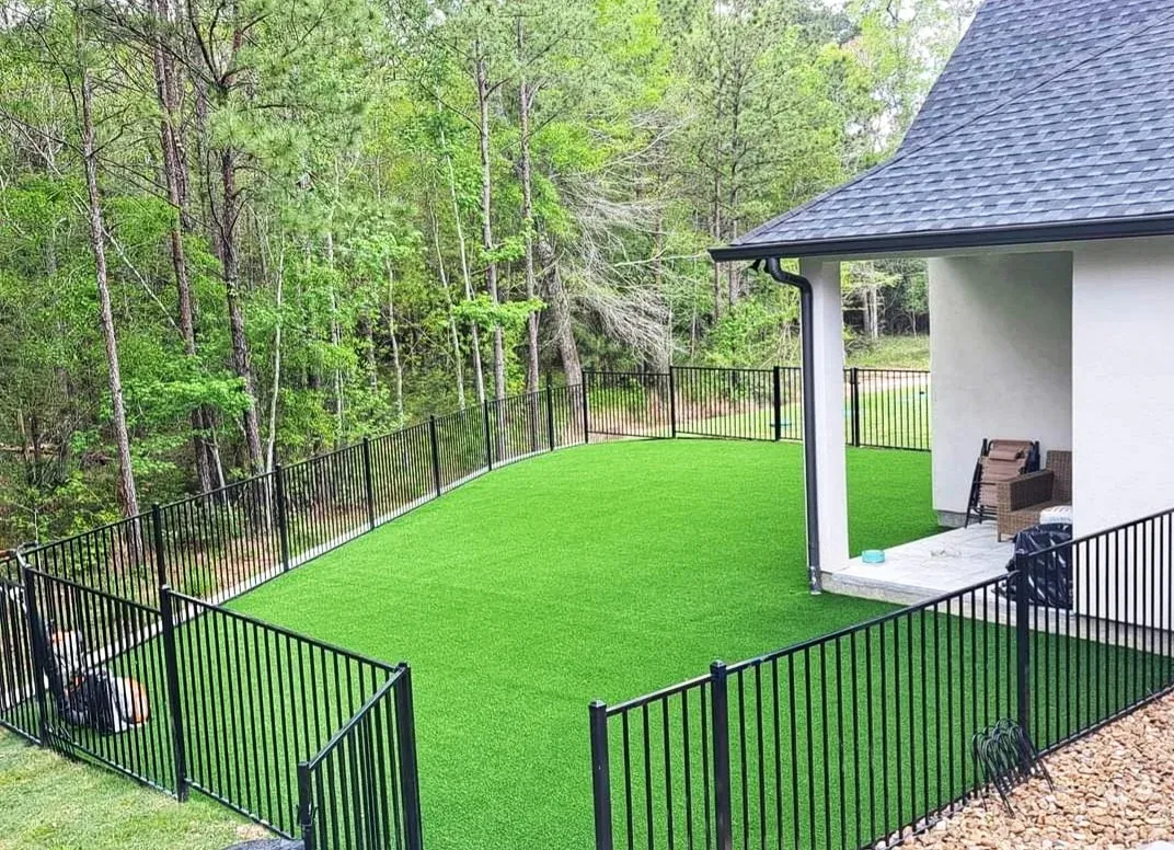 A fenced backyard laid in artificial turf in front of a covered porch, wooded lot behind