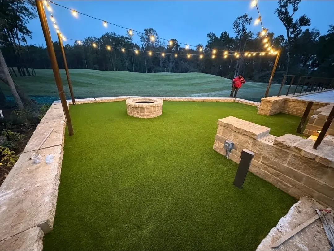 An artificial turf terrace around a stone fire pit, lit by festoon lighting at dusk