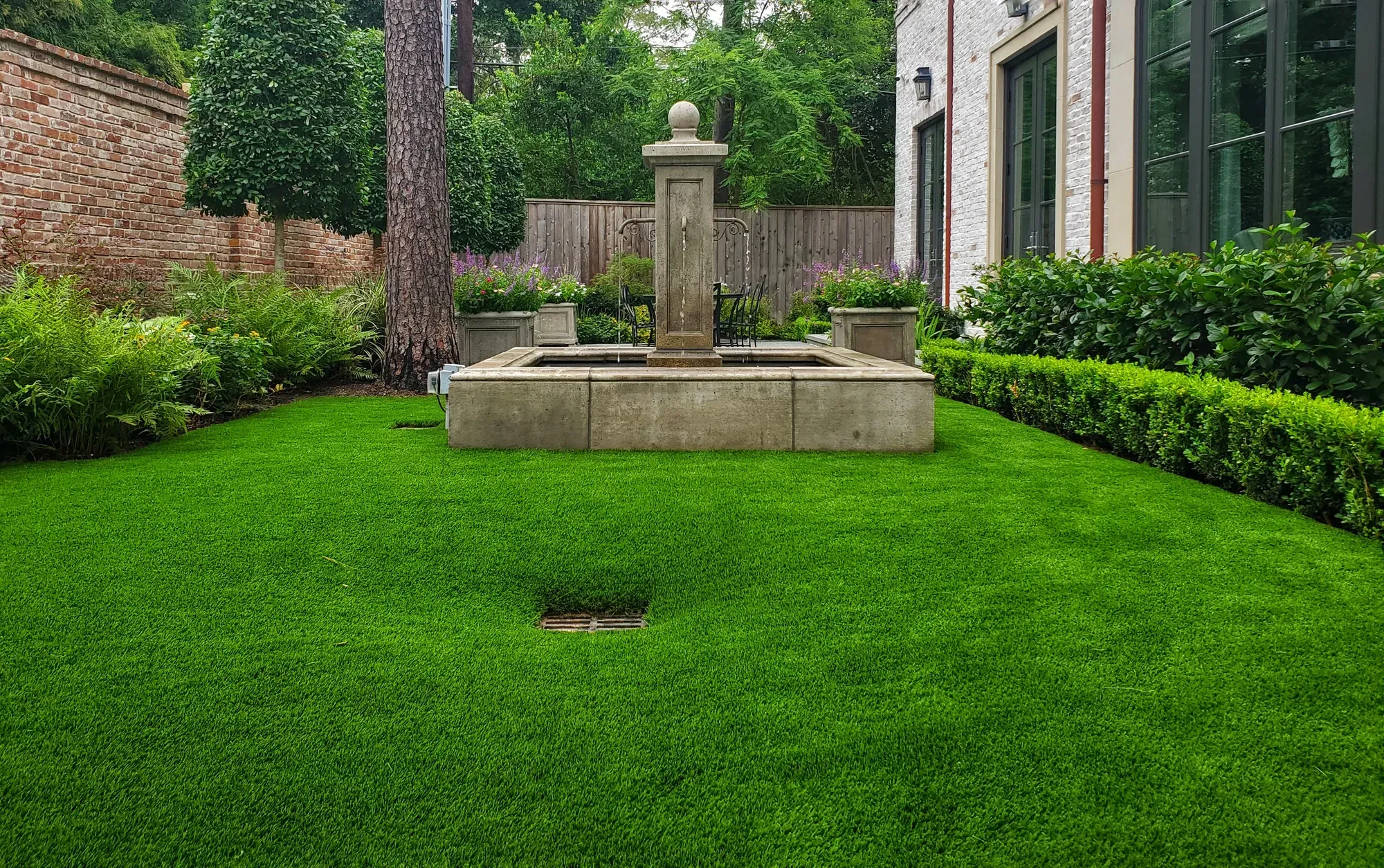 A clipped, hedge-lined lawn of artificial turf around a weathered stone fountain
