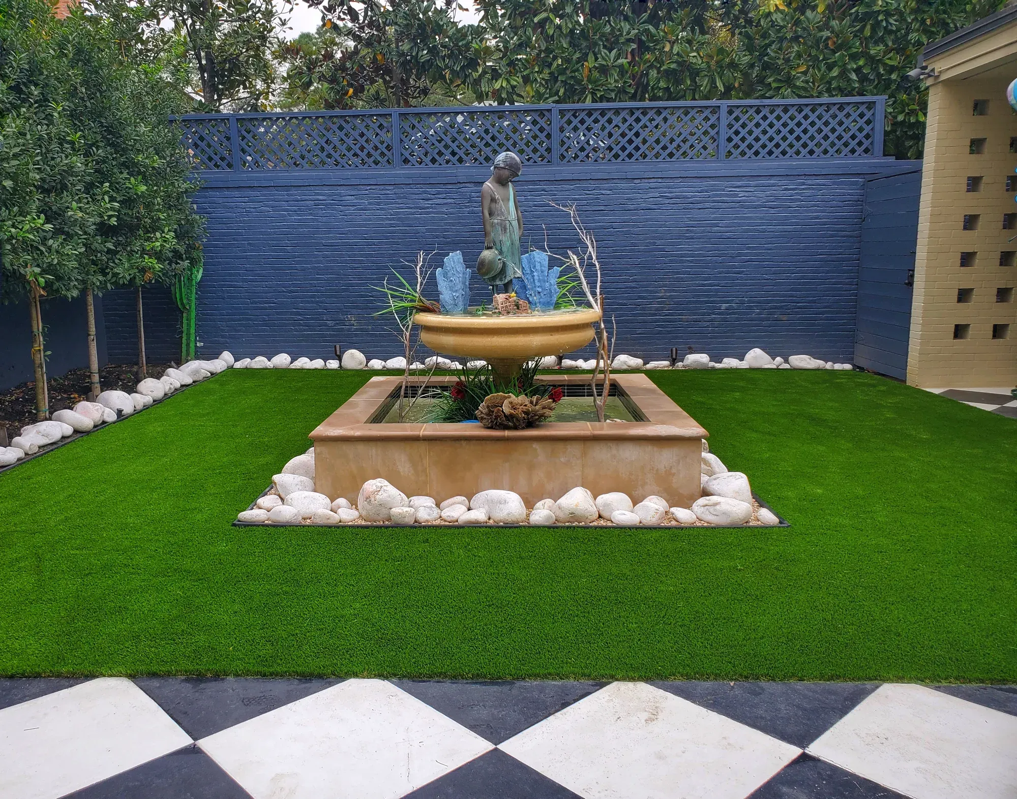 A stone fountain on a chequerboard apron, surrounded by a clipped artificial turf lawn