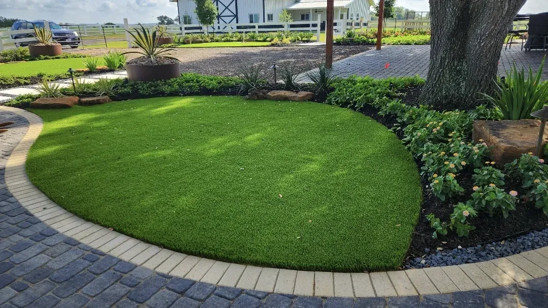 A circular artificial lawn edged with a paver band, framed by planting beds and mature trees