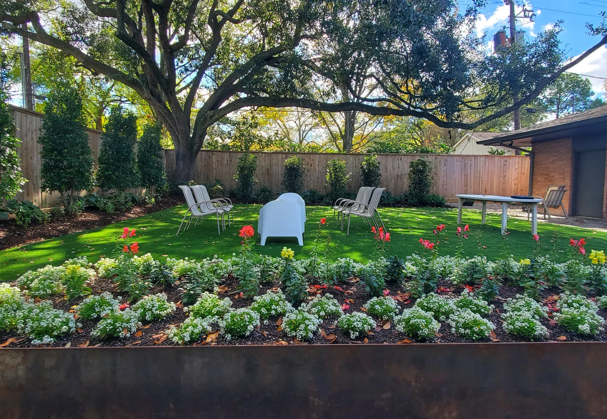 Artificial turf lawn under a spreading live oak behind a steel-edged bed of white and red flowers