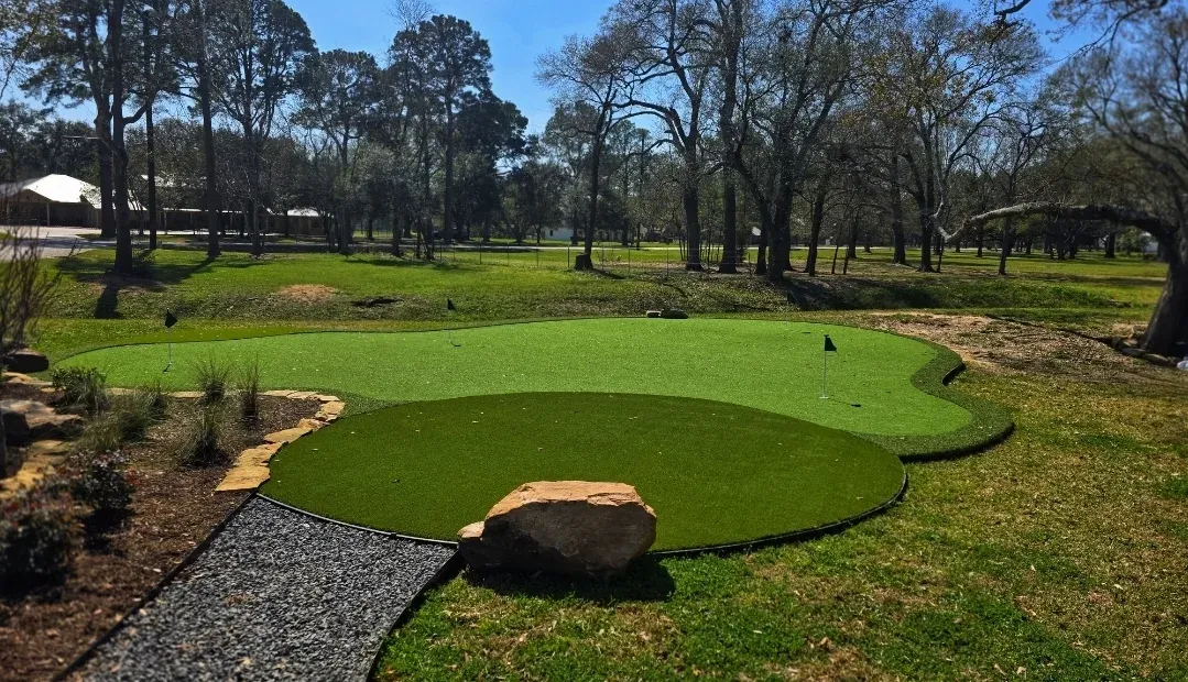 A curved backyard practice green with flags, set into a turf lawn under mature trees