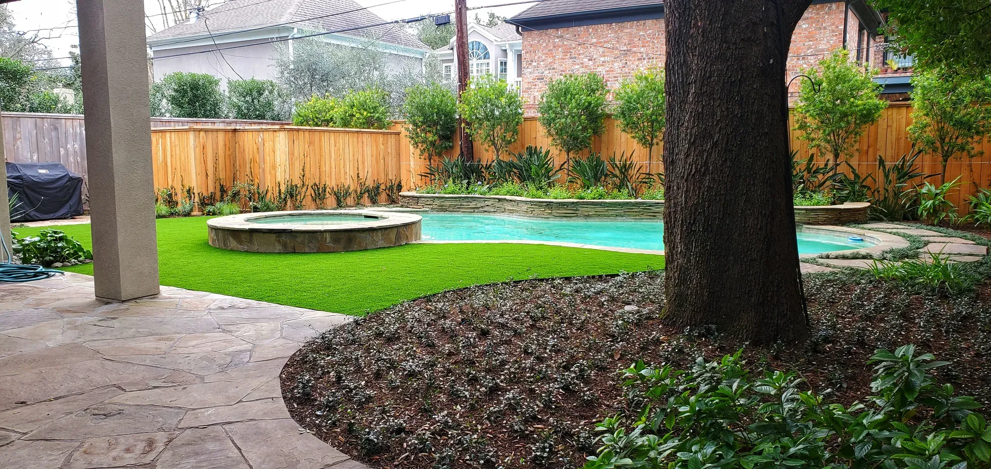 A poolside artificial turf lawn laid around the trunk of a large mature tree, with a raised fire feature