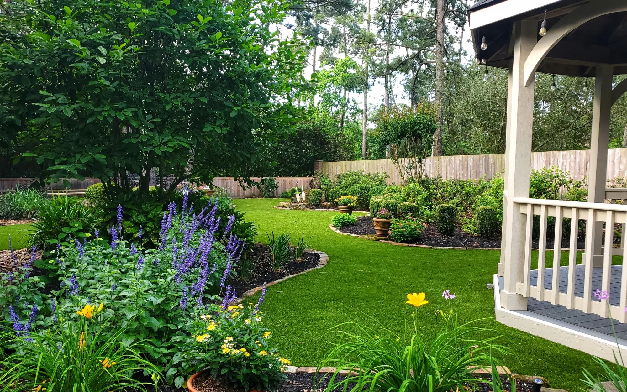 An artificial turf lawn curving between deep flowering borders, with a white gazebo at one side