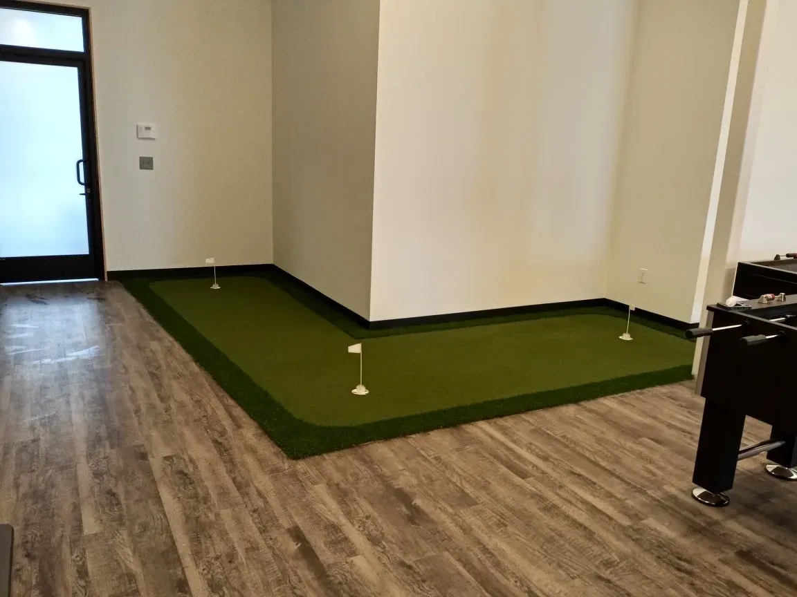 Low-pile artificial turf laid as flooring inside a room, finishing flush against plank flooring, with a glazed door beyond