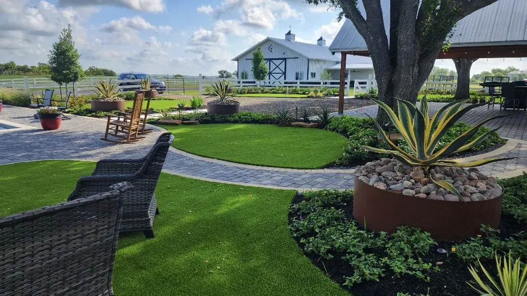 A wicker chair on an artificial lawn beside a planted container, barn buildings beyond