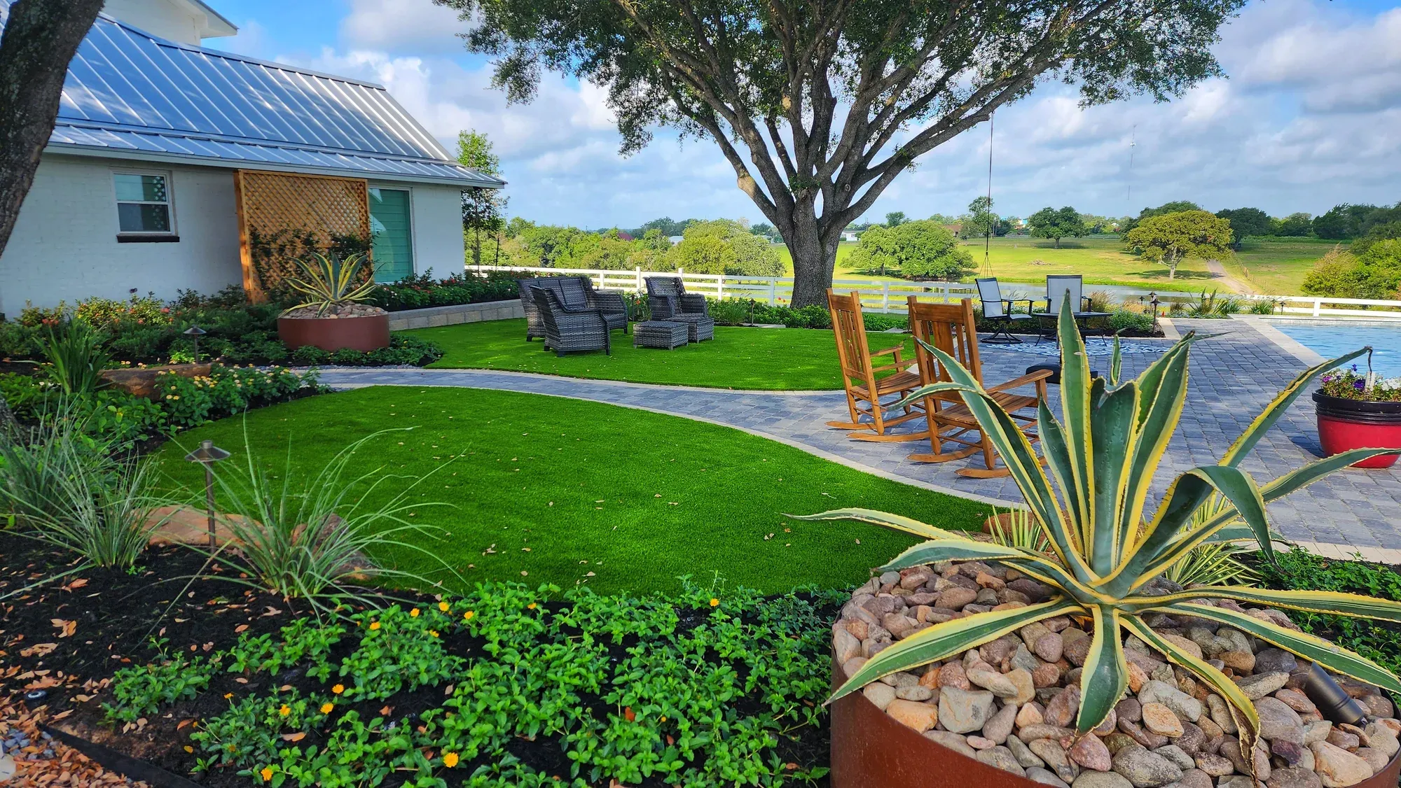 Artificial turf lawns and paver walkways around a lakeside house, with a live oak and open water behind