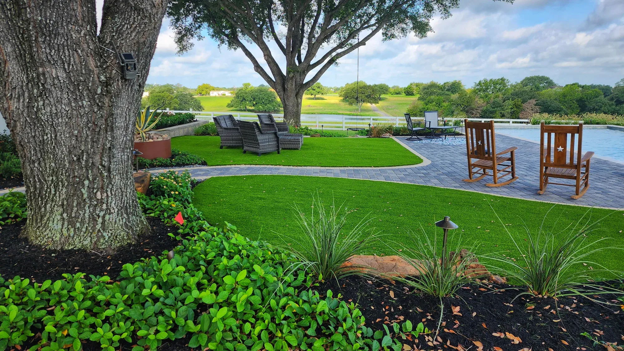 A curved artificial turf lawn under mature trees, with lounge seating and a lake beyond