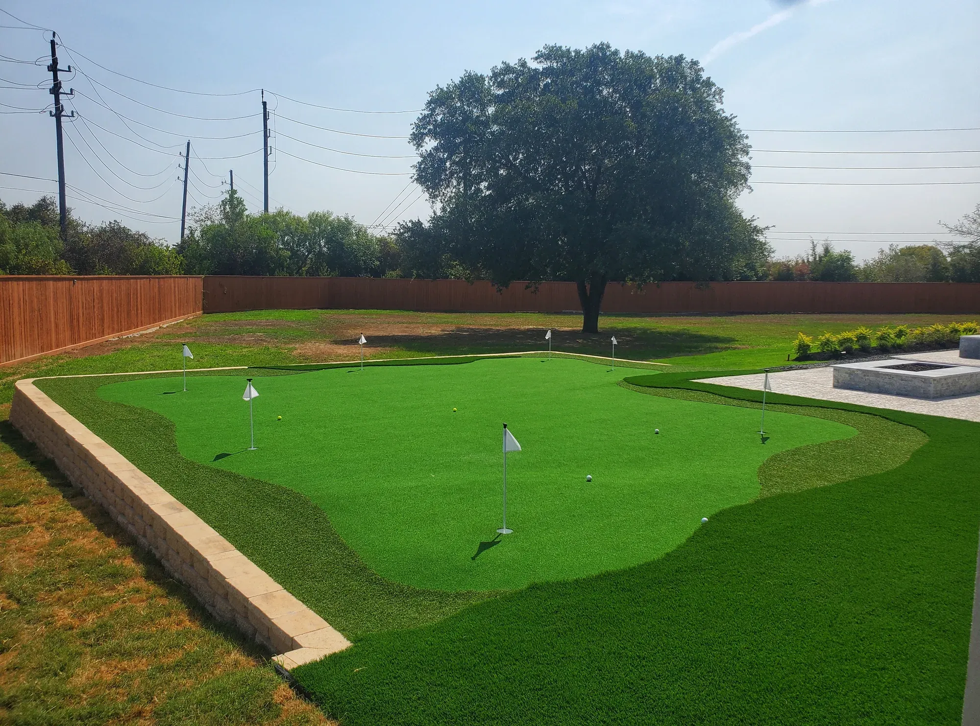 Large artificial putting green with six flagsticks, built out from a stone retaining wall behind a fenced yard