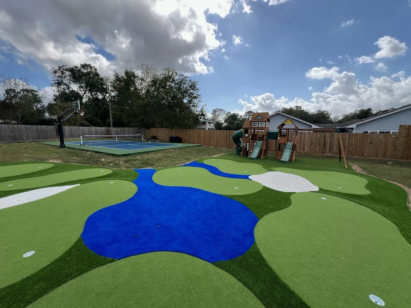 Multi-hole miniature golf course in shaped artificial turf, with a blue turf water hazard and white turf bunkers, a sport court and playset behind it