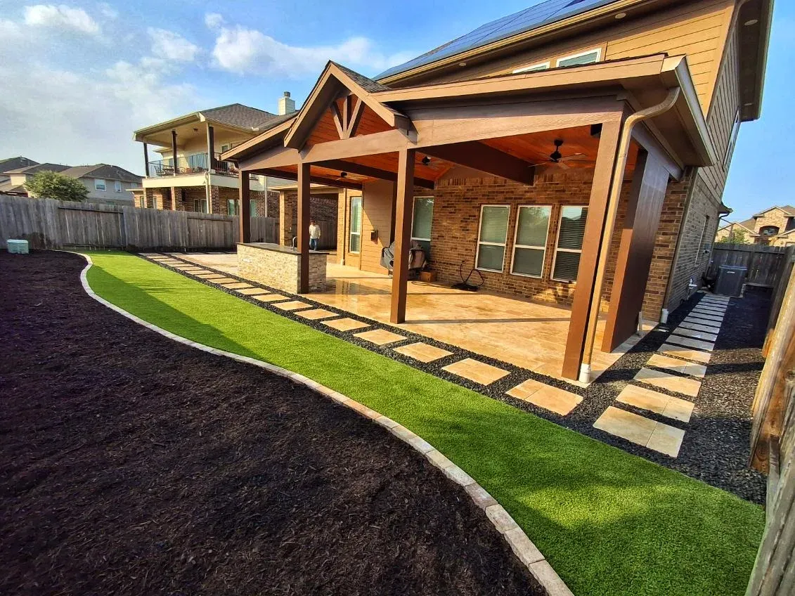 A stone terrace with artificial turf joints beneath a timber pergola, beside freshly mulched planting beds