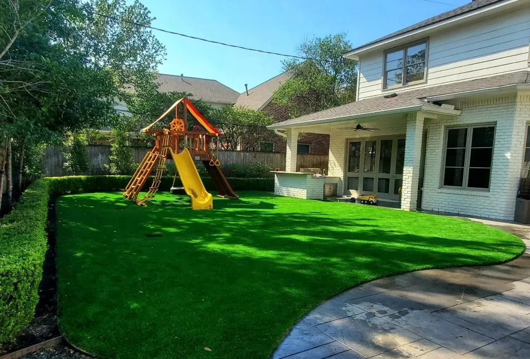 Artificial turf play area running up to a wooden playset with a yellow slide, edged by a fence and a covered patio