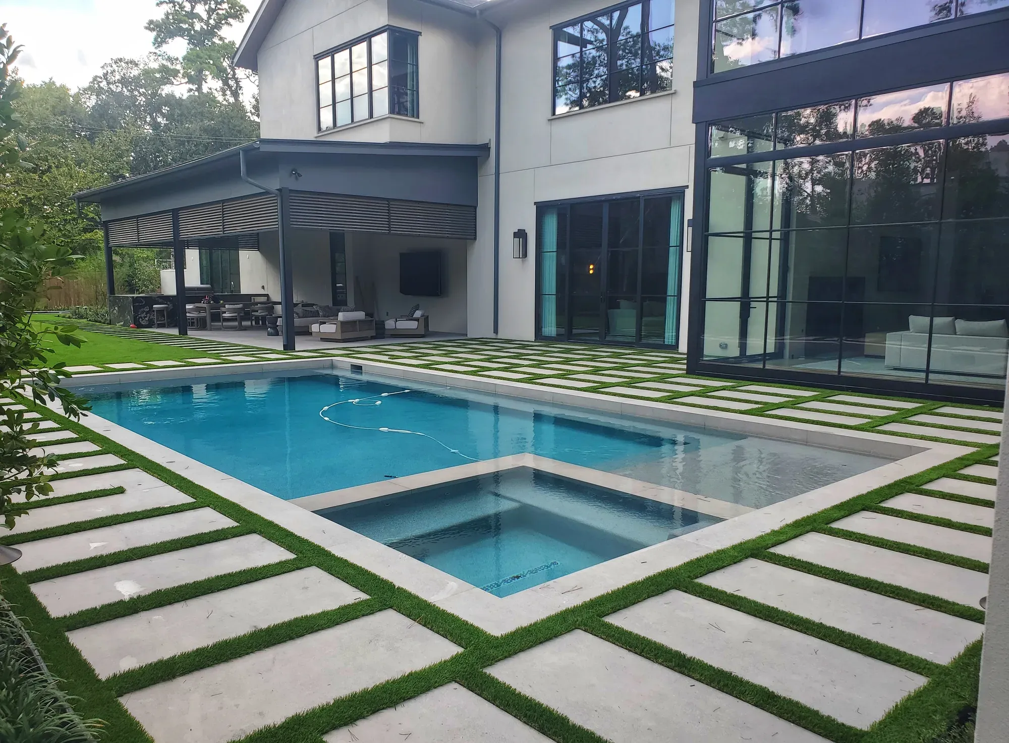 A paver-and-turf grid wrapping a pool and spa behind a modern house