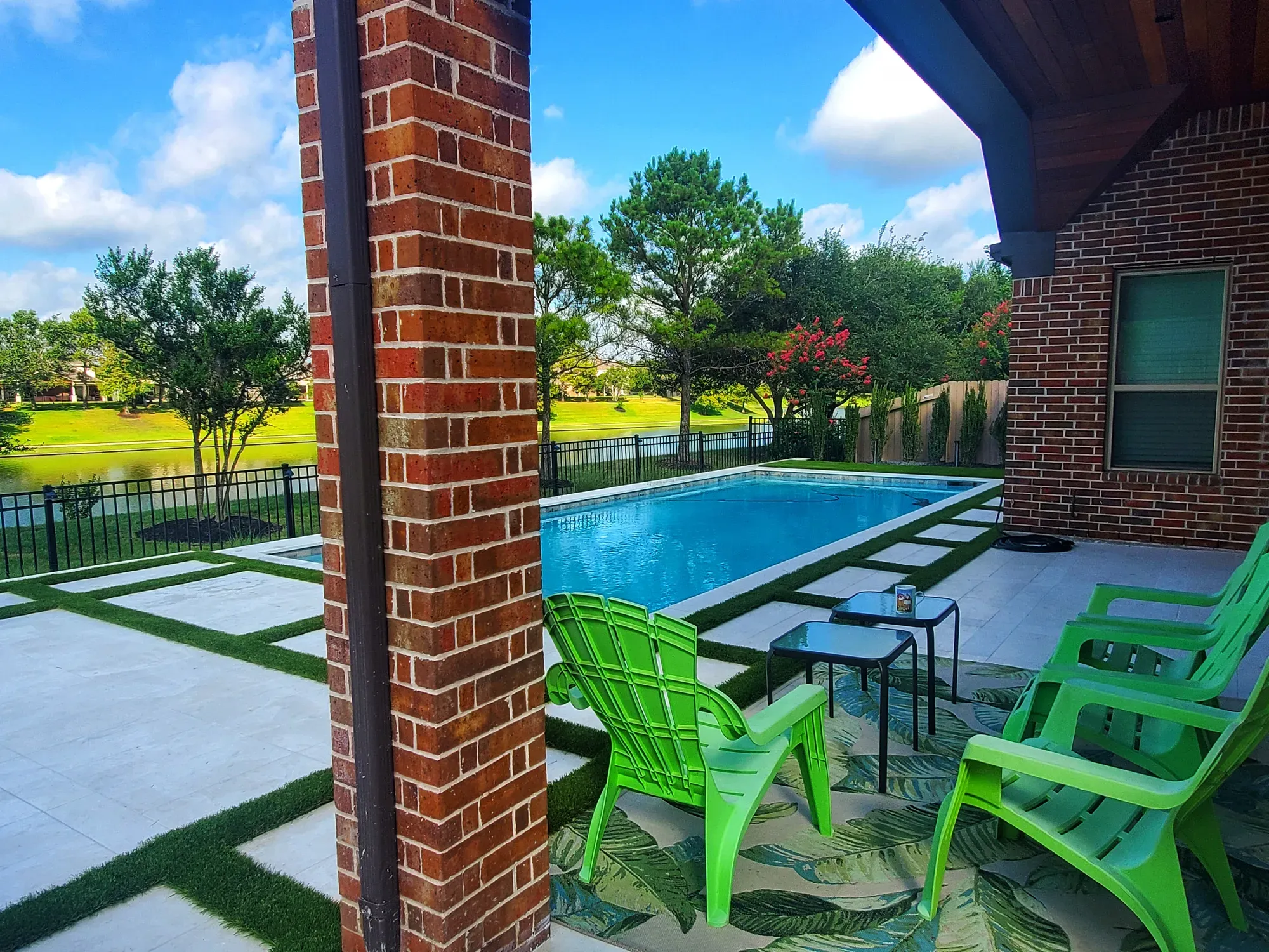 A pool deck of large pavers with turf joints, seen past a brick column with green chairs beyond