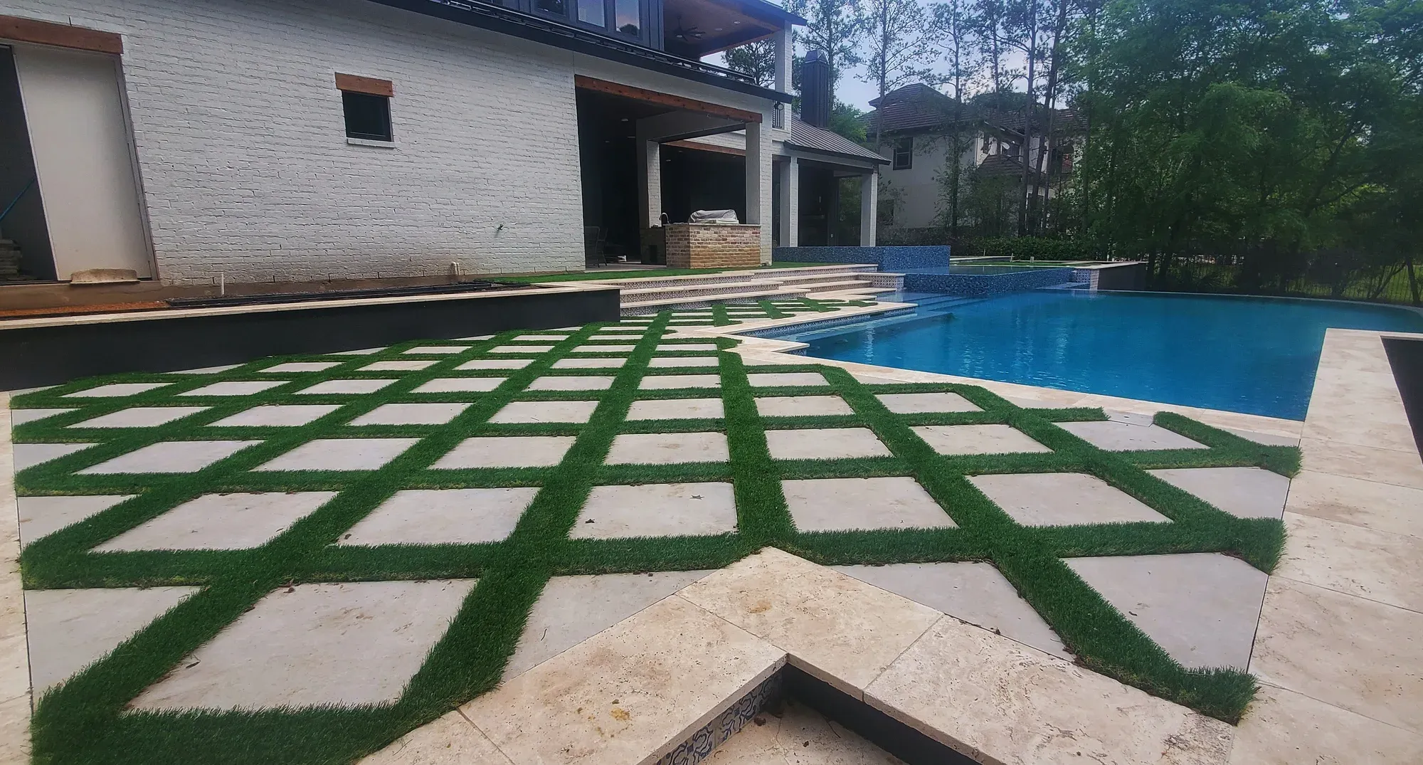 A wide paver-and-turf grid across a pool deck under tall pines
