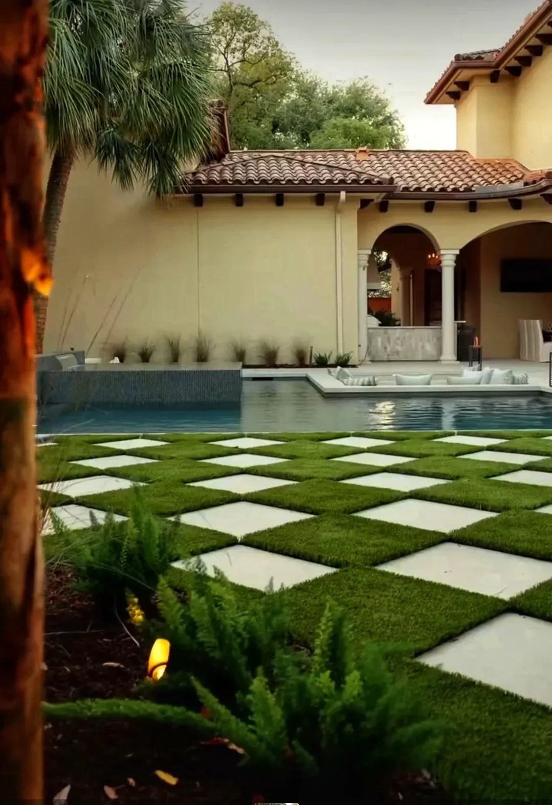 A chequerboard pool deck of alternating pale stone slabs and artificial turf squares, lit at dusk