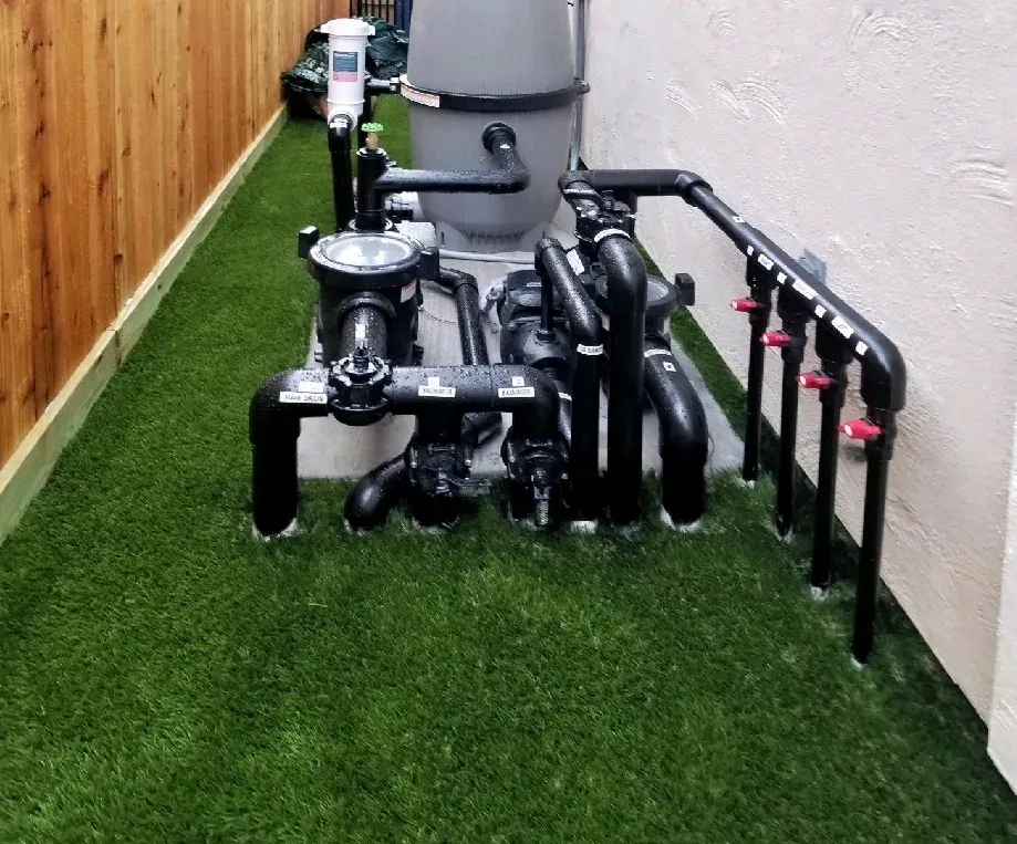 A pool equipment manifold and filter standing on artificial turf against a timber fence