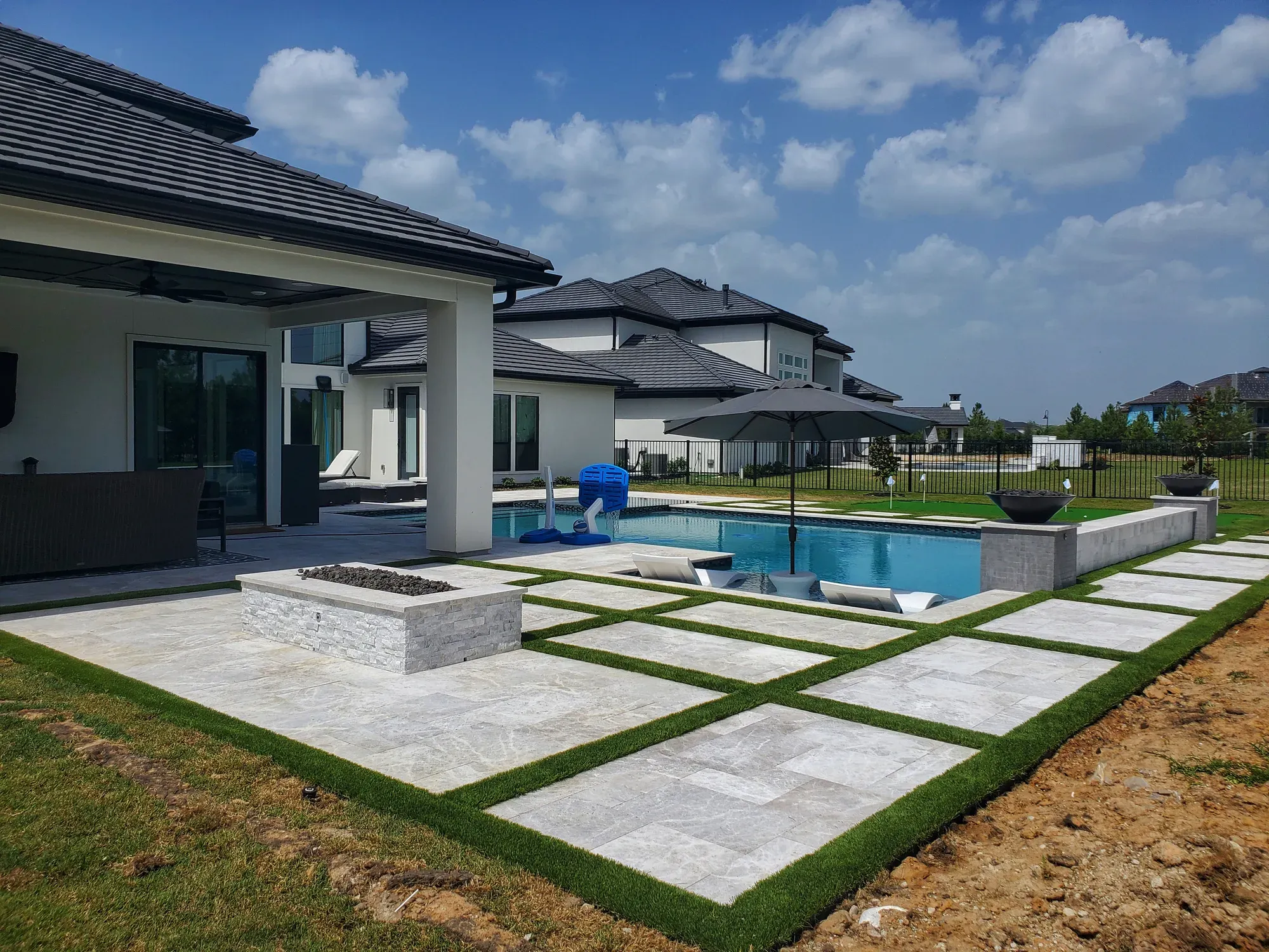 A pool deck of large pavers with turf strips between them, fire feature and covered patio behind