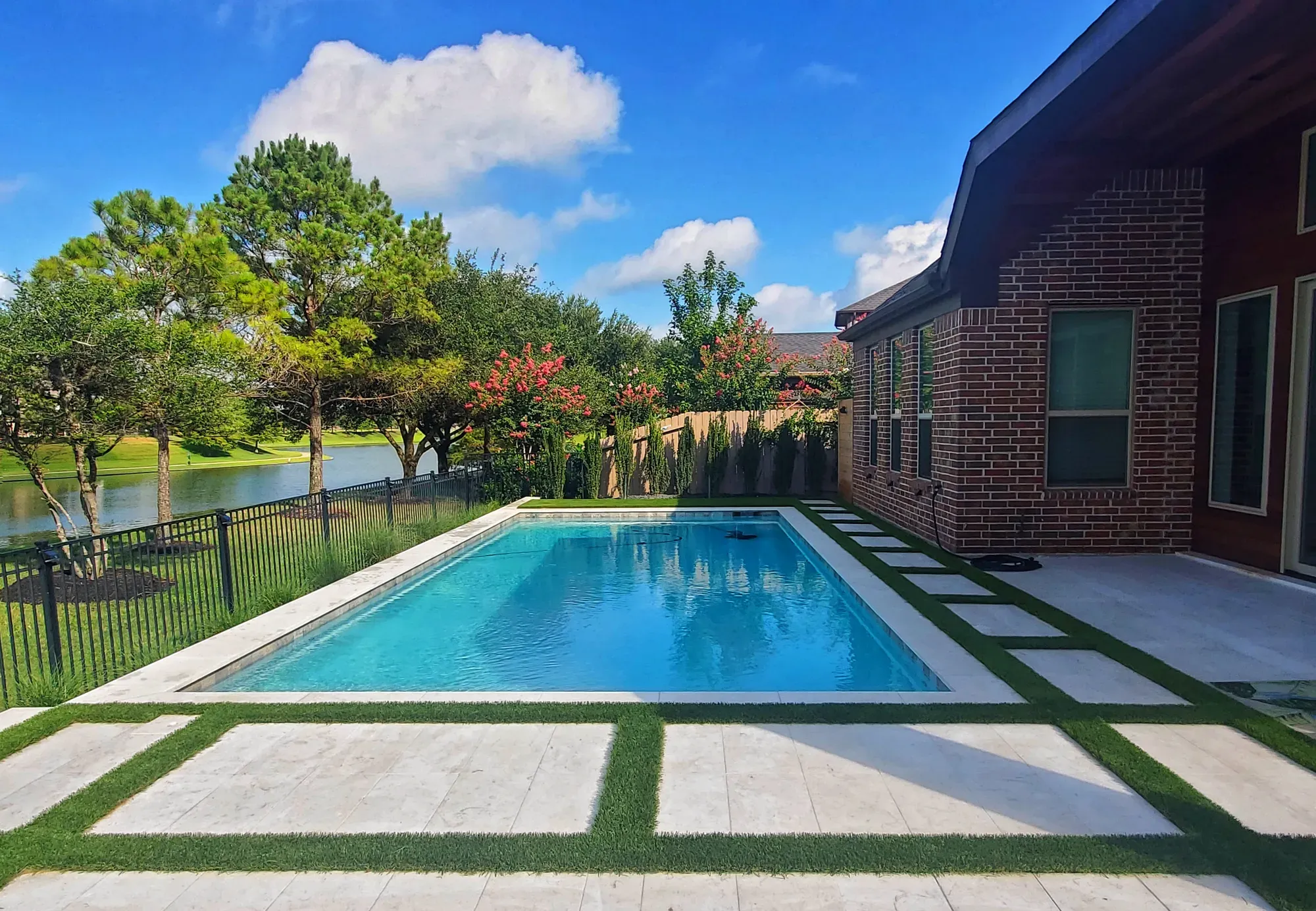 Travertine pool deck laid as large pavers with artificial turf strips running between them, beside a rectangular pool and a lake
