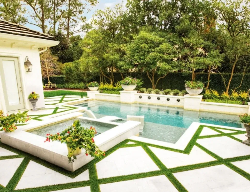 A diagonal lattice of artificial turf inlays set into a pale stone pool deck around a pool and spa