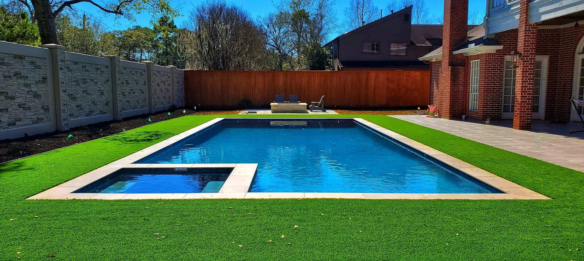 An artificial turf lawn wrapping the full perimeter of a rectangular pool between a brick house and a timber fence